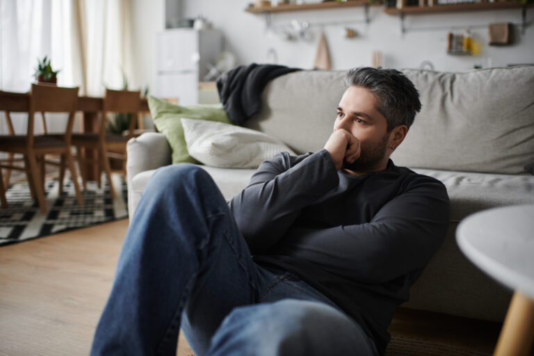 A handsome man sits on the floor, lost in thought, surrounded by a comfortable living area.