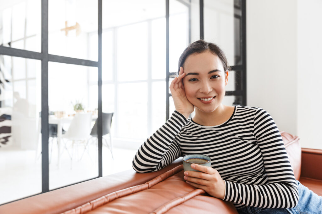Attractive smiling young asian woman wearing casual clothes relaxing on a leather couch at home, having cup of coffee
